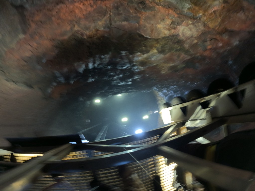A view inside a dimly lit mine shaft, showcasing rough rock walls with some illumination from overhead lights and a metal track system.