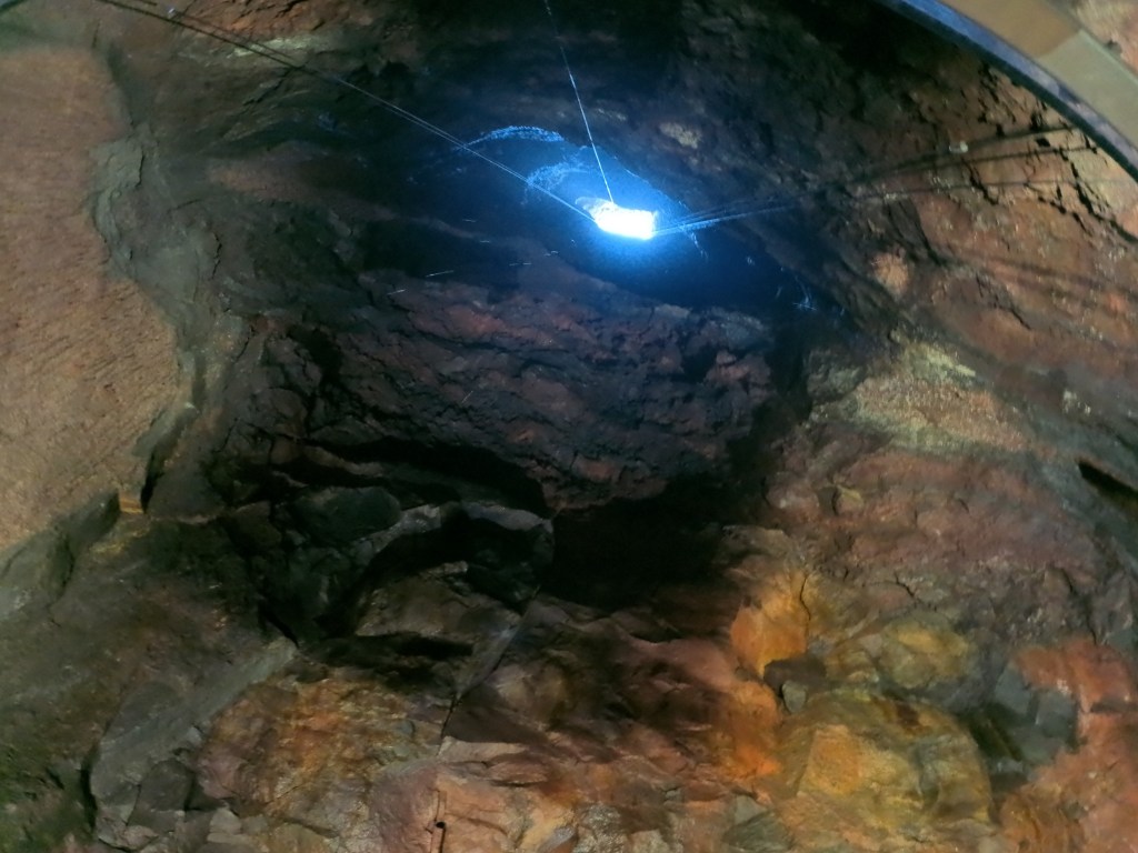 Interior view of a deep rock formation with a bright light shining from above, highlighting various textures and colors of the rocks.