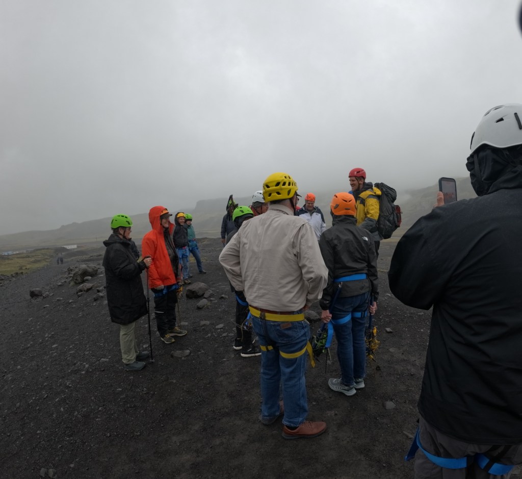 A group of people in helmets and safety gear are gathered on rocky terrain, discussing instructions for an outdoor adventure, likely ice climbing or glacier hiking.