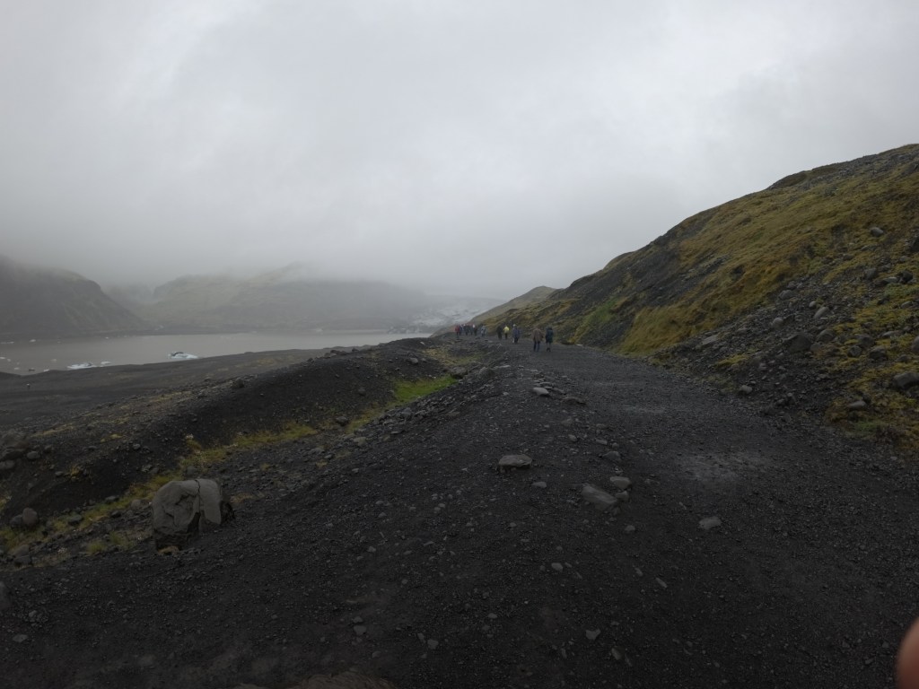View of a gravel path leading through rugged terrain, with a group of hikers walking in the distance under cloudy skies.