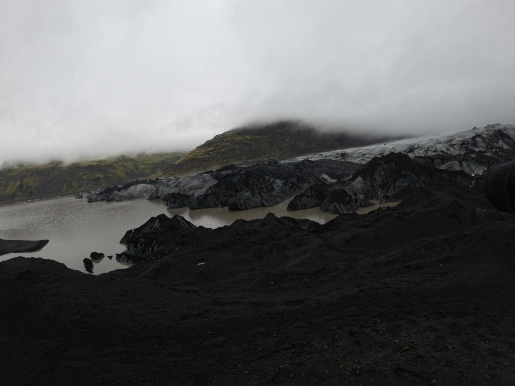 A panoramic view of a dark volcanic landscape with a glacier and a cloudy sky over a lake.