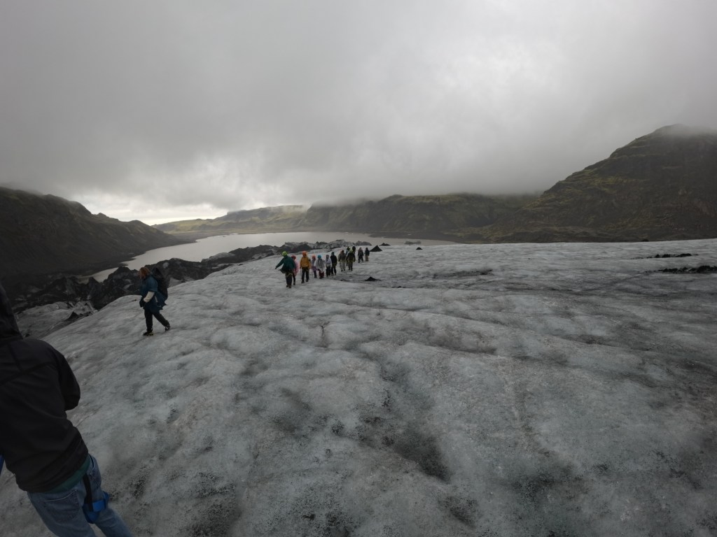 A group of hikers walking on a glacier with mountains and a lake in the background under cloudy skies.
