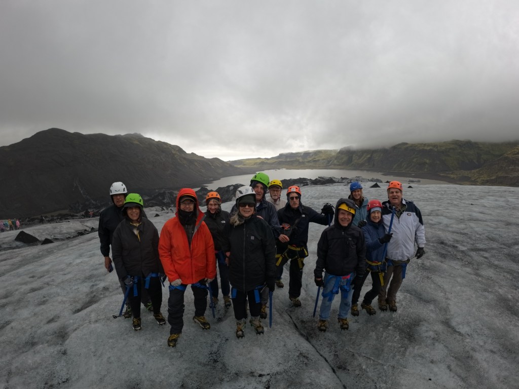Group of people standing on a glacier, wearing helmets and harnesses, with mountains and cloudy sky in the background.