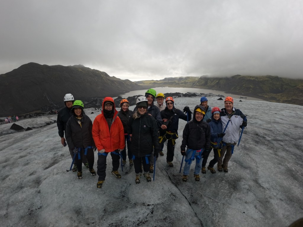 A group of climbers wearing helmets and harnesses pose on a glacier, with a mountainous landscape and a lake in the background.