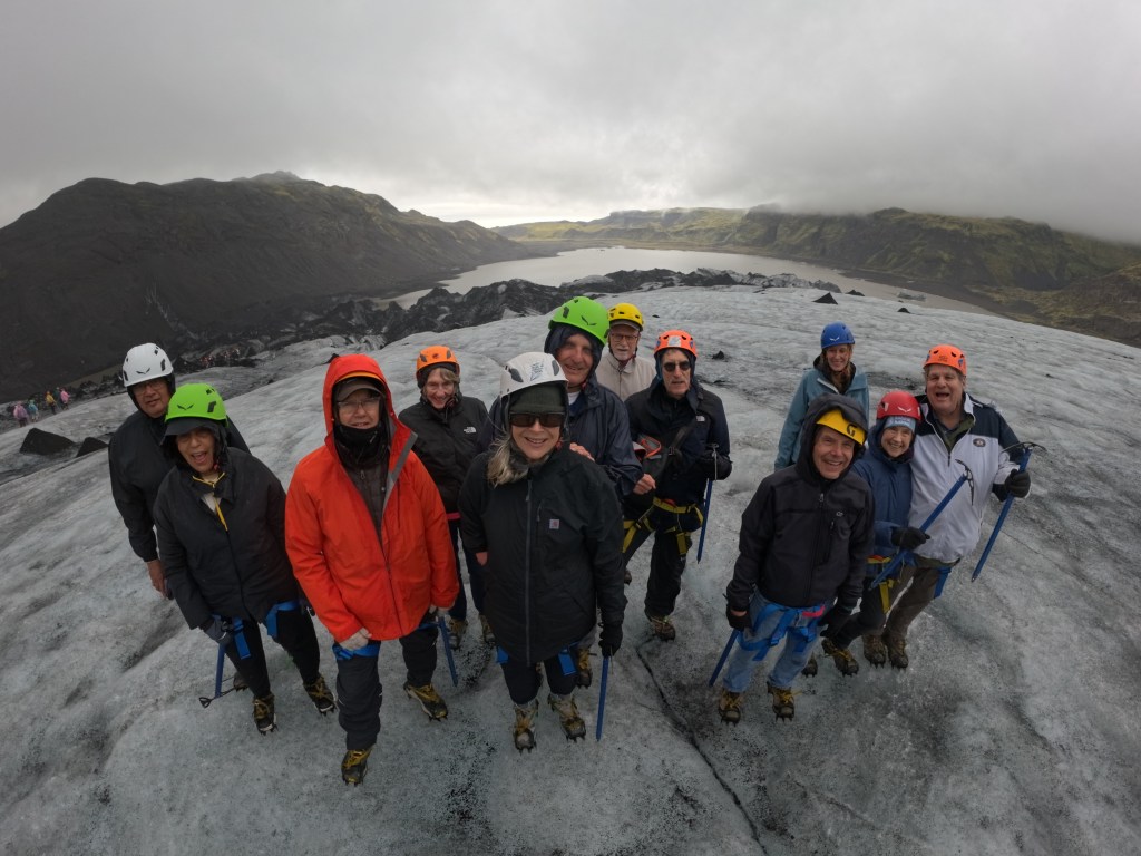 A group of hikers on a glacier, wearing helmets and ice climbing gear, posing for a photo with a scenic valley and cloudy sky in the background.