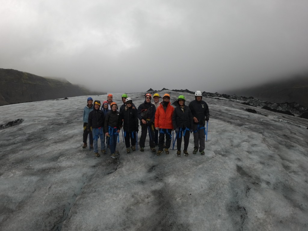 A group of climbers standing on a glacier, wearing helmets and outdoor gear, with cloudy mountains in the background.