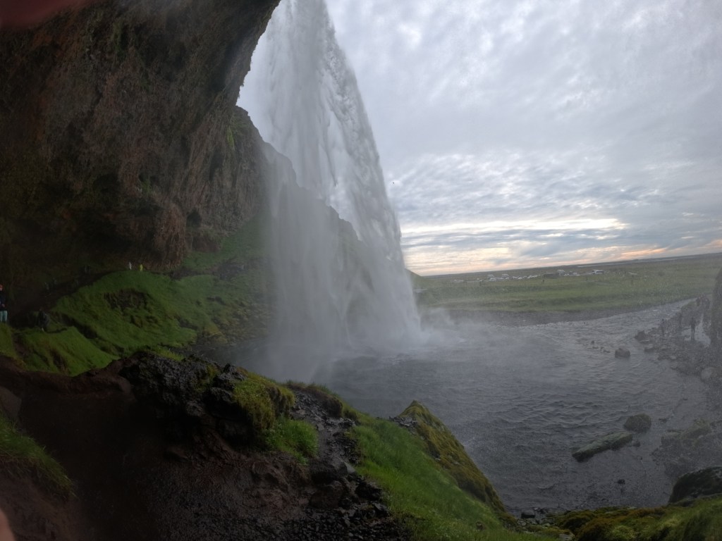 A scenic view of a waterfall cascading over rocky cliffs, with lush green grass in the foreground and a cloudy sky in the background.