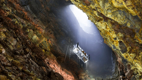 A deep mine shaft illuminated by a beam of light, featuring a suspended platform surrounded by rocky walls.