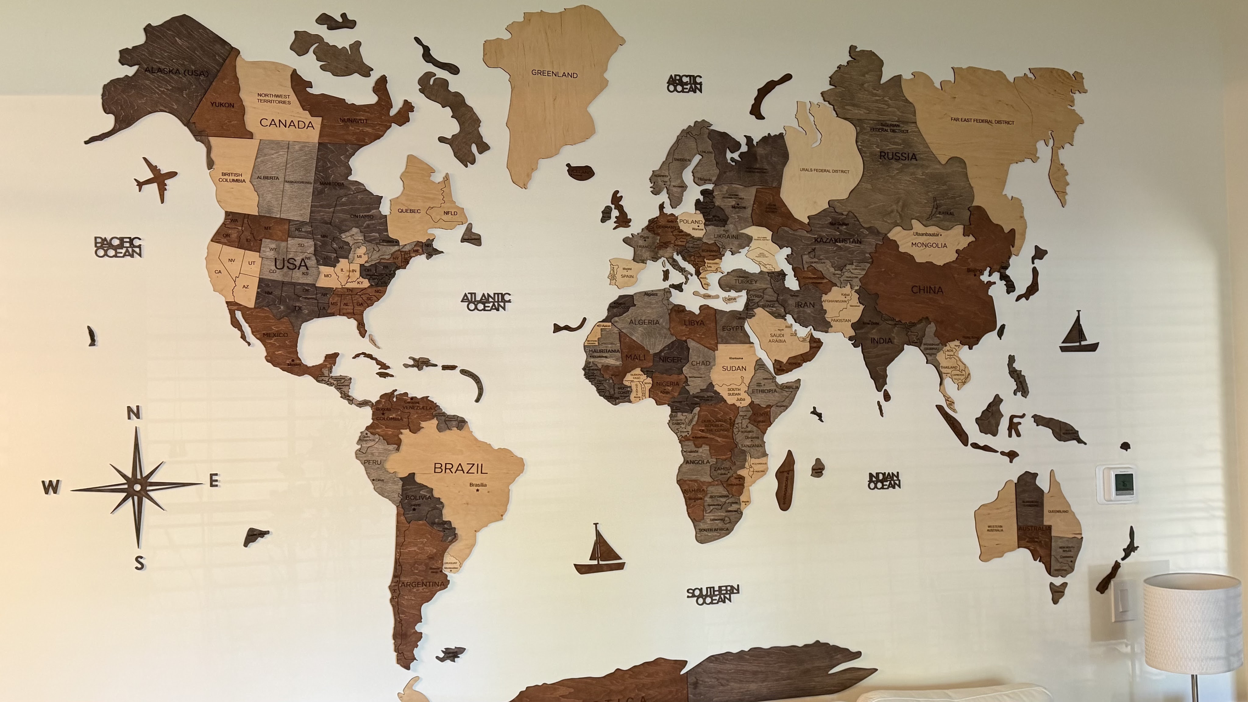 A detailed wooden map of the world displayed on a wall, showcasing countries and oceans with varying wood tones. Features labels for countries like Canada, USA, Brazil, China, and others, along with decorative elements like airplanes and boats.
