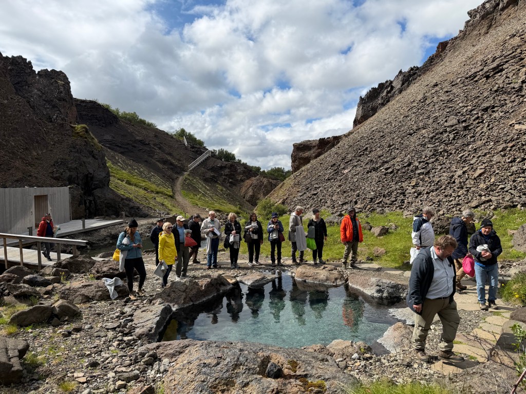 A group of people gathered near a natural hot spring surrounded by rocky terrain and lush greenery, with some walking along a path and others standing by the water.