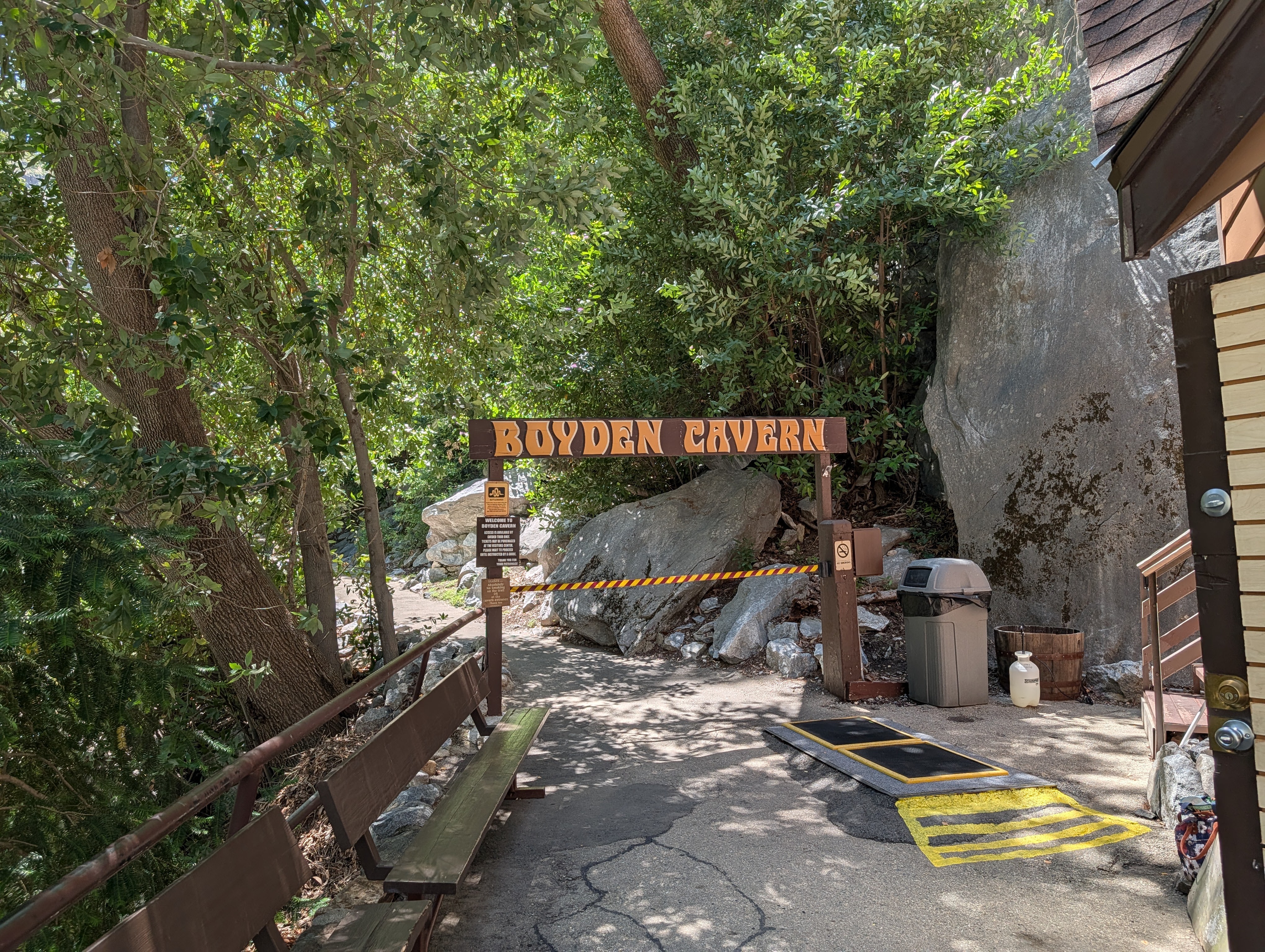 Entrance sign for Boyden Cavern surrounded by greenery and rocky terrain, with benches and a trash can nearby.