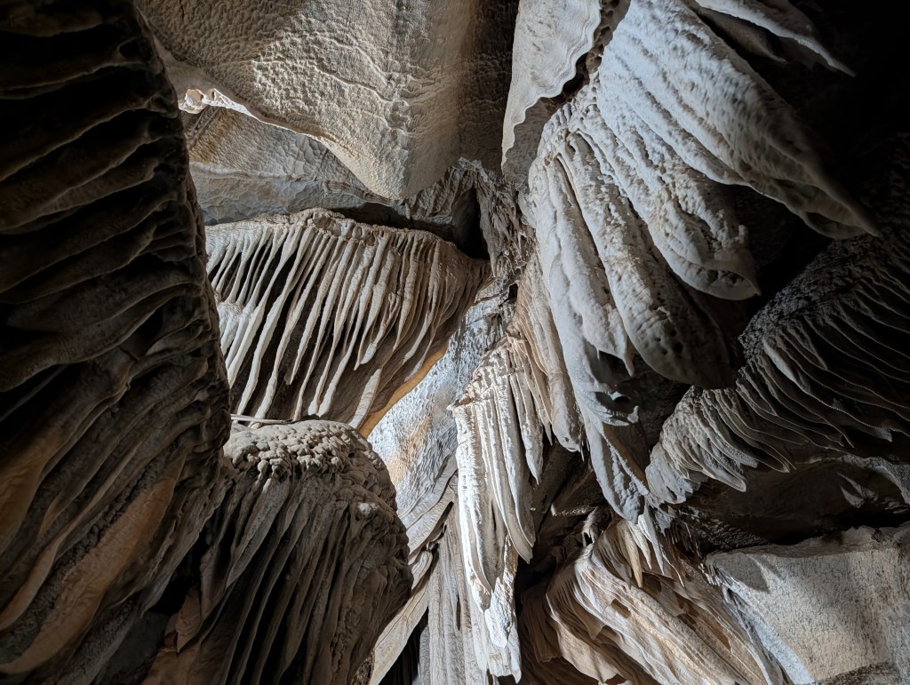Interior view of a cave showing intricate rock formations with layered, textured structures and soft lighting illuminating the scene.