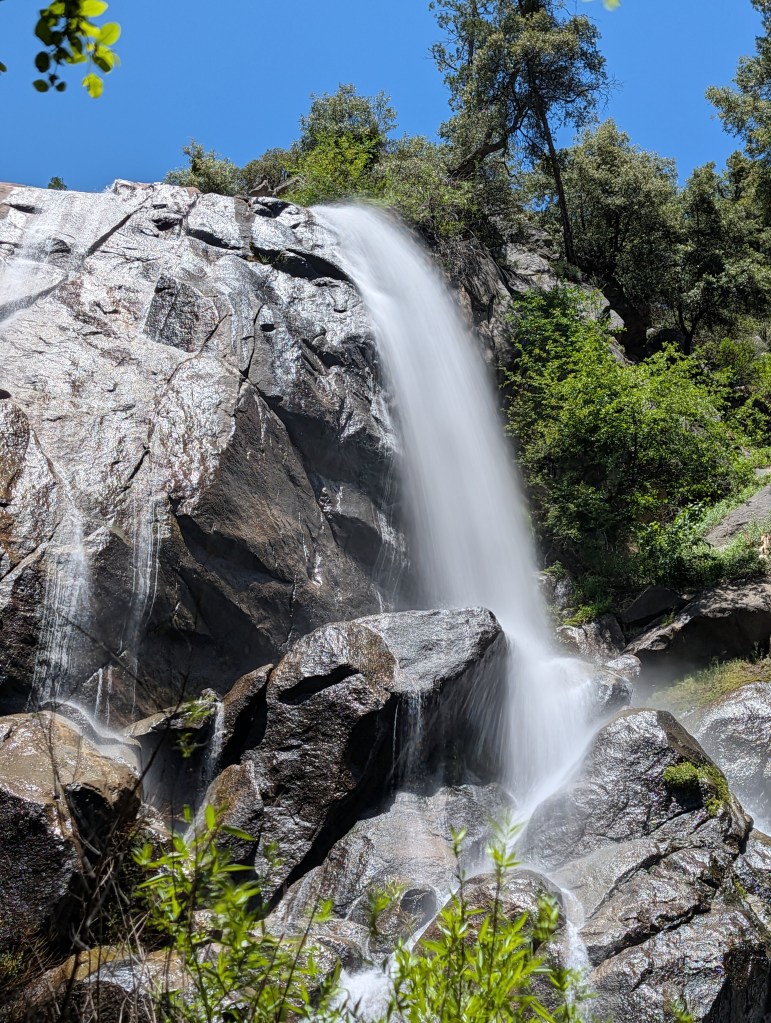 A cascading waterfall flowing over rocky terrain, surrounded by green foliage and under a clear blue sky.