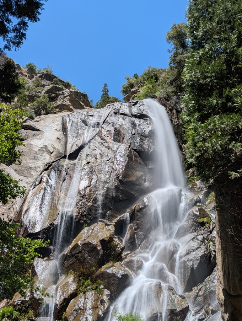 A waterfall cascading down rocky cliffs, surrounded by lush greenery and a clear blue sky.