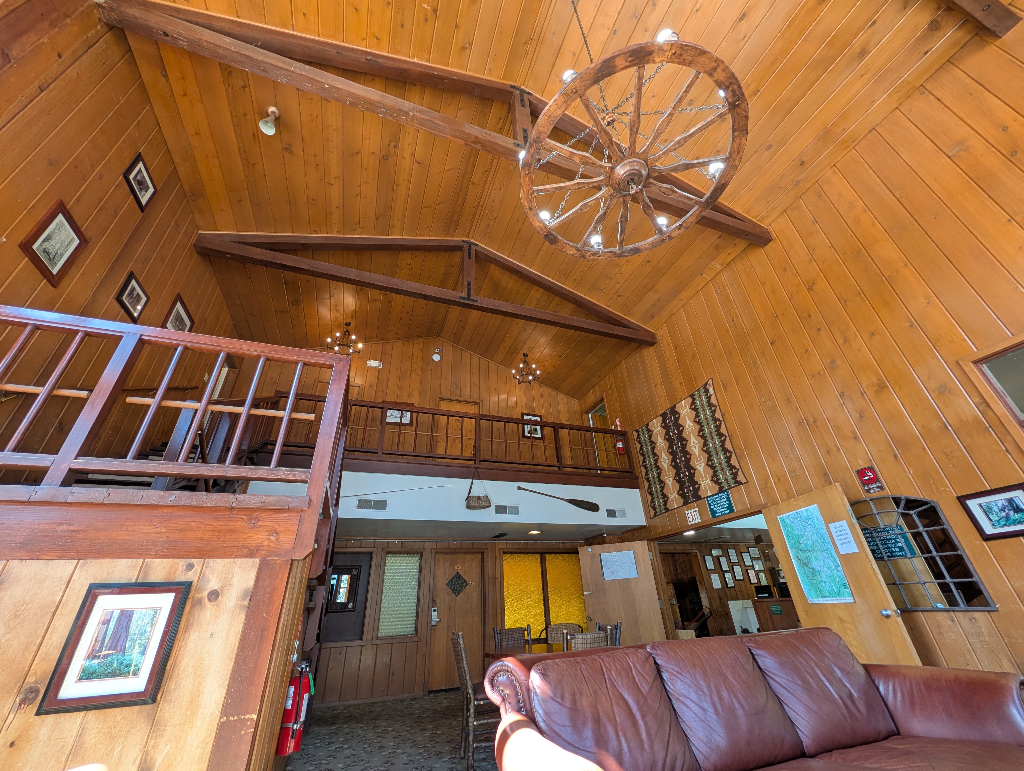Interior of a wooden cabin-style lodge, featuring high ceilings with a decorative chandelier, wooden paneling, and a staircase leading to an upper level with framed images on the walls.
