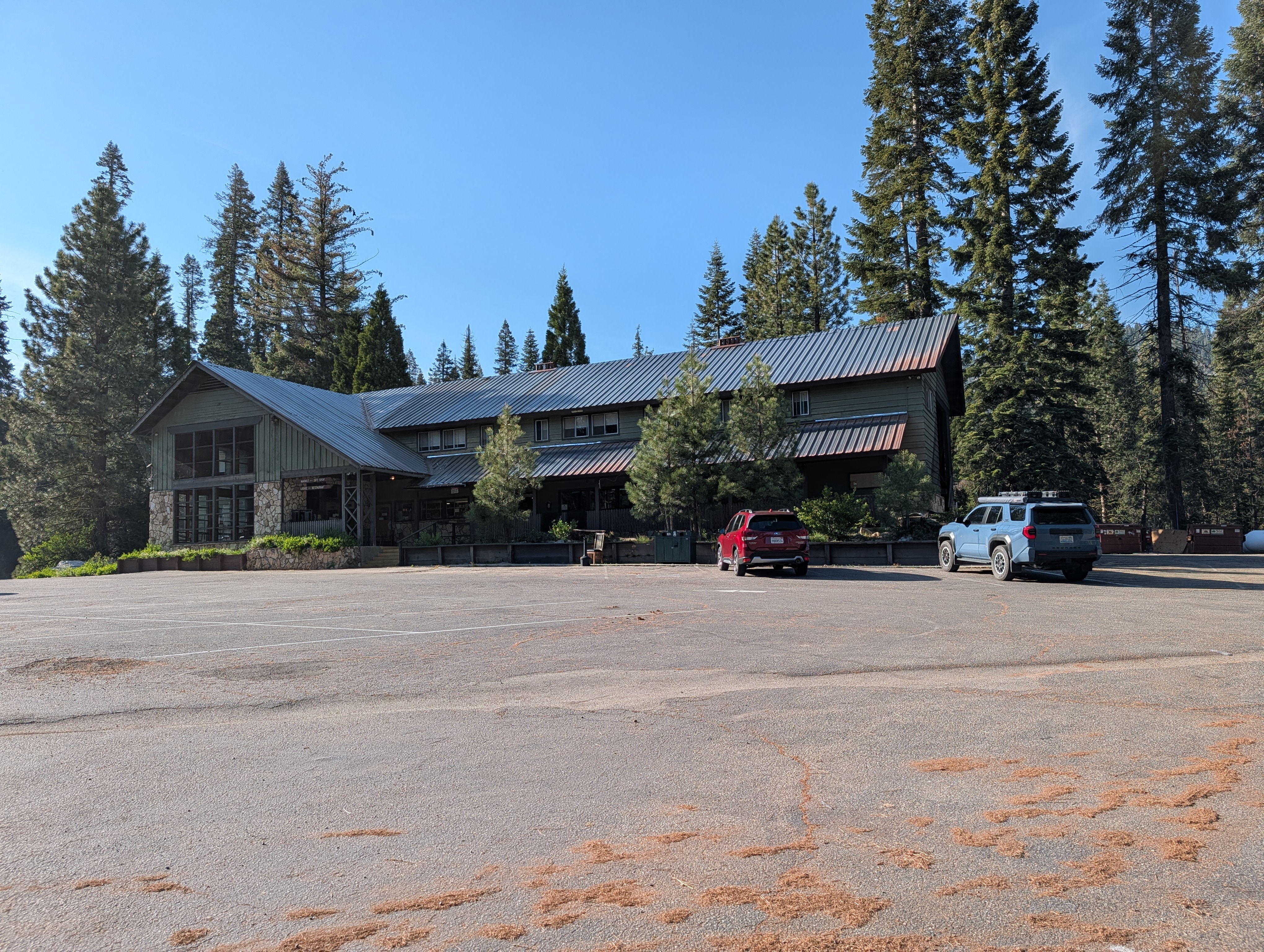 A rustic building surrounded by tall pine trees, with a gravel parking lot in the foreground and two parked vehicles, one red and one blue.