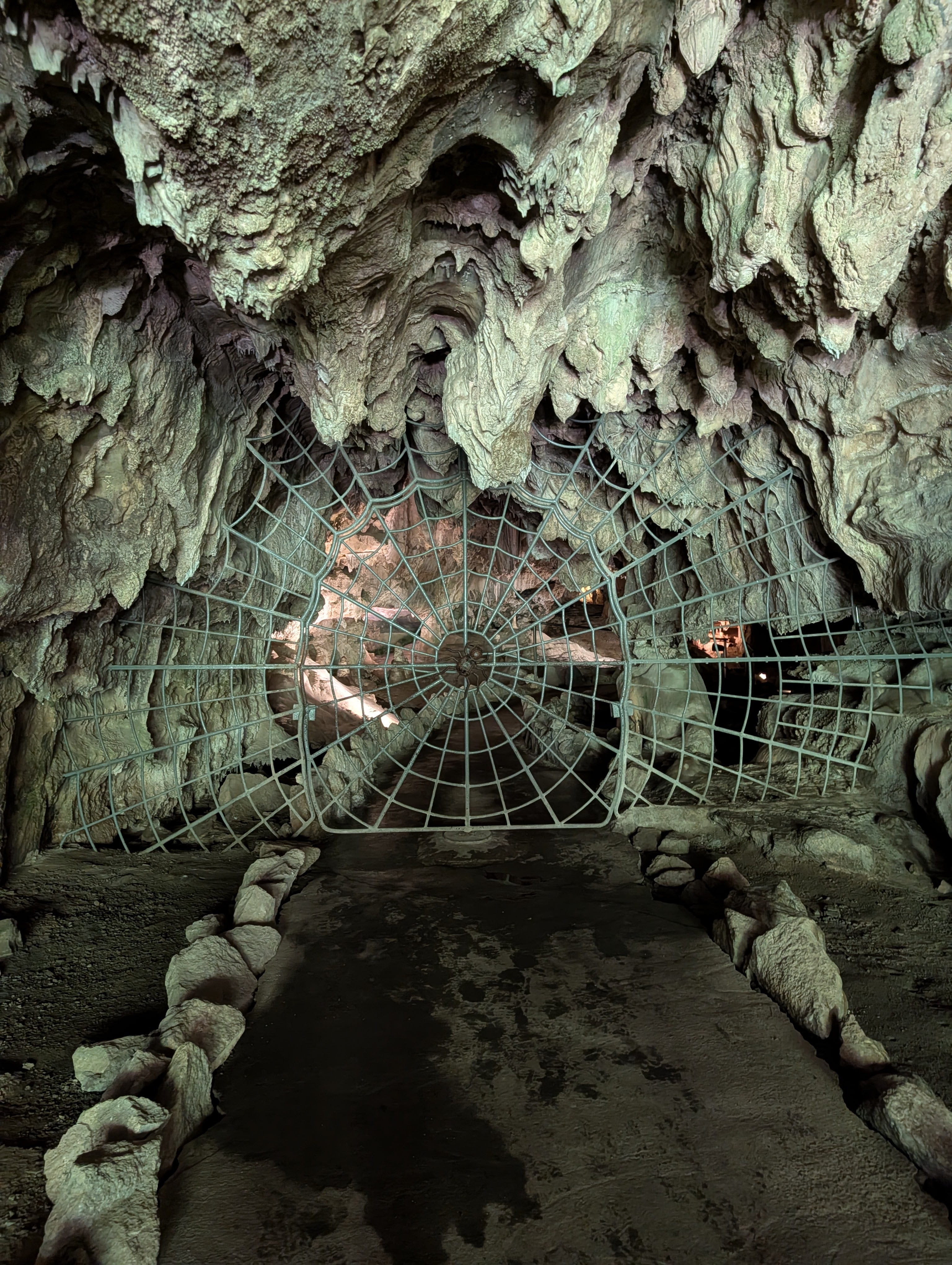An underground cave featuring textured rock formations and a spider web-shaped metal gate at the entrance.