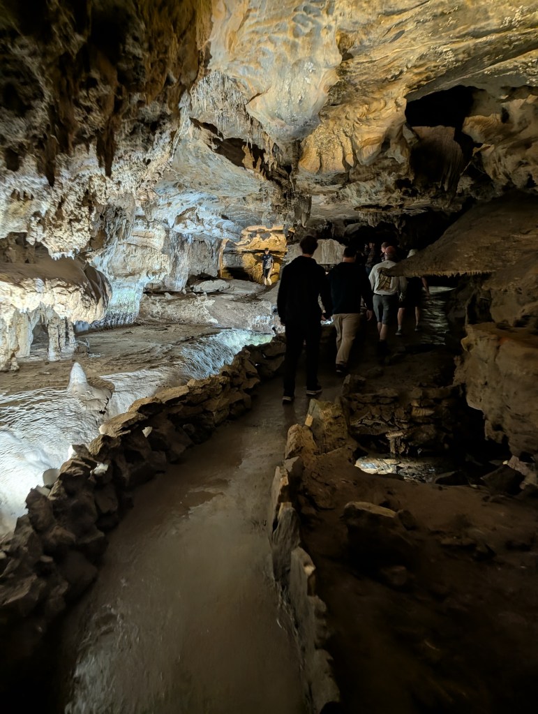 Inside a cave with rock formations and illuminated pathways, people walking along a stone-bordered trail.
