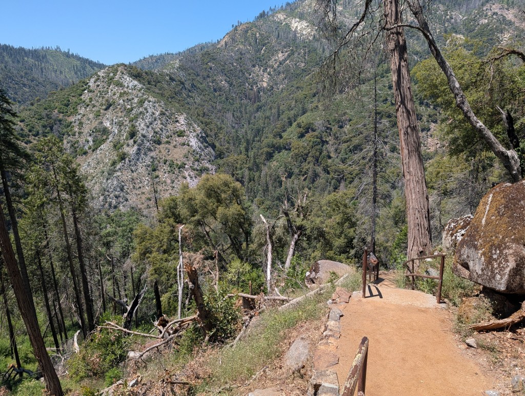 A dirt path leads through a forested area with tall trees and rocky terrain, offering scenic views of surrounding hills and valleys.