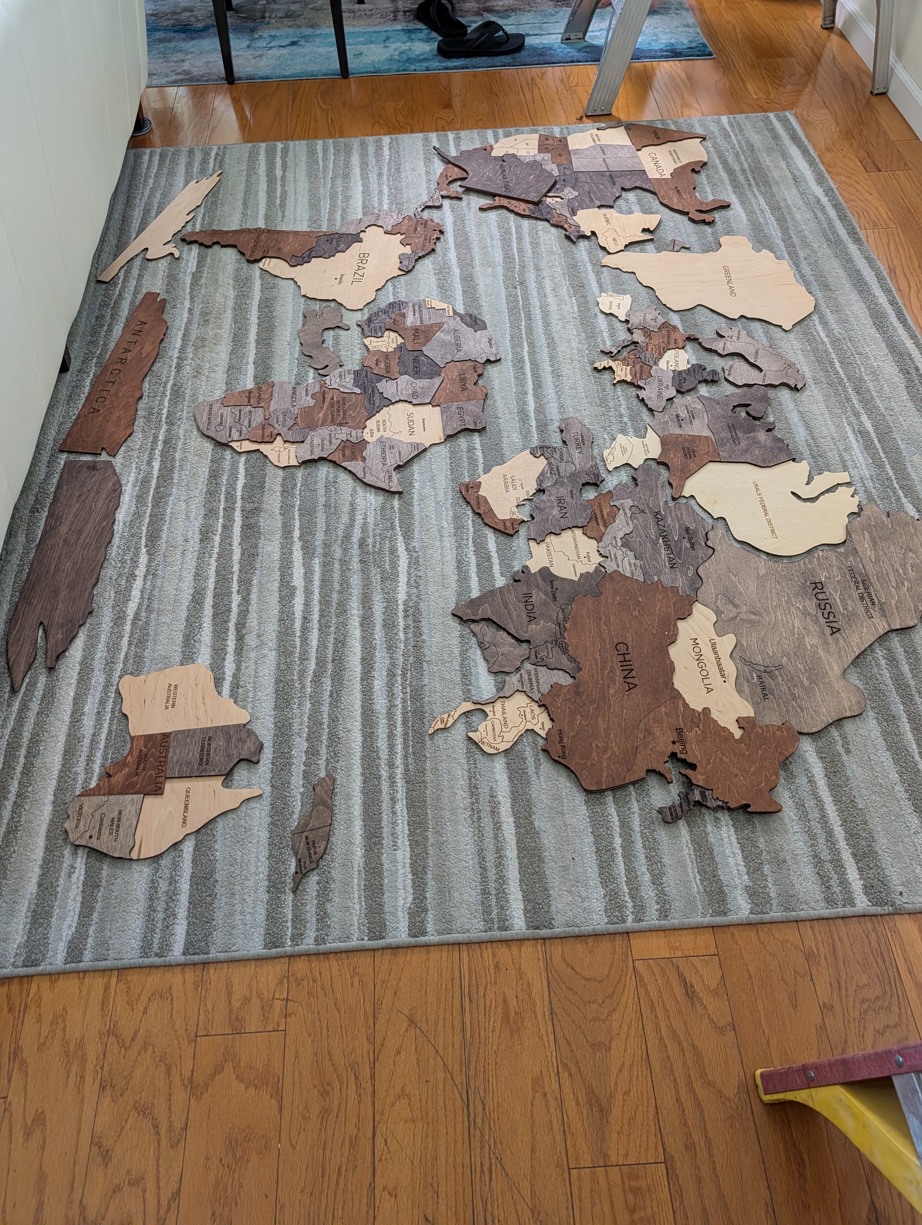 A wooden puzzle map of the world laid out on a striped rug, featuring labeled countries in different shades of brown and light-colored wood.