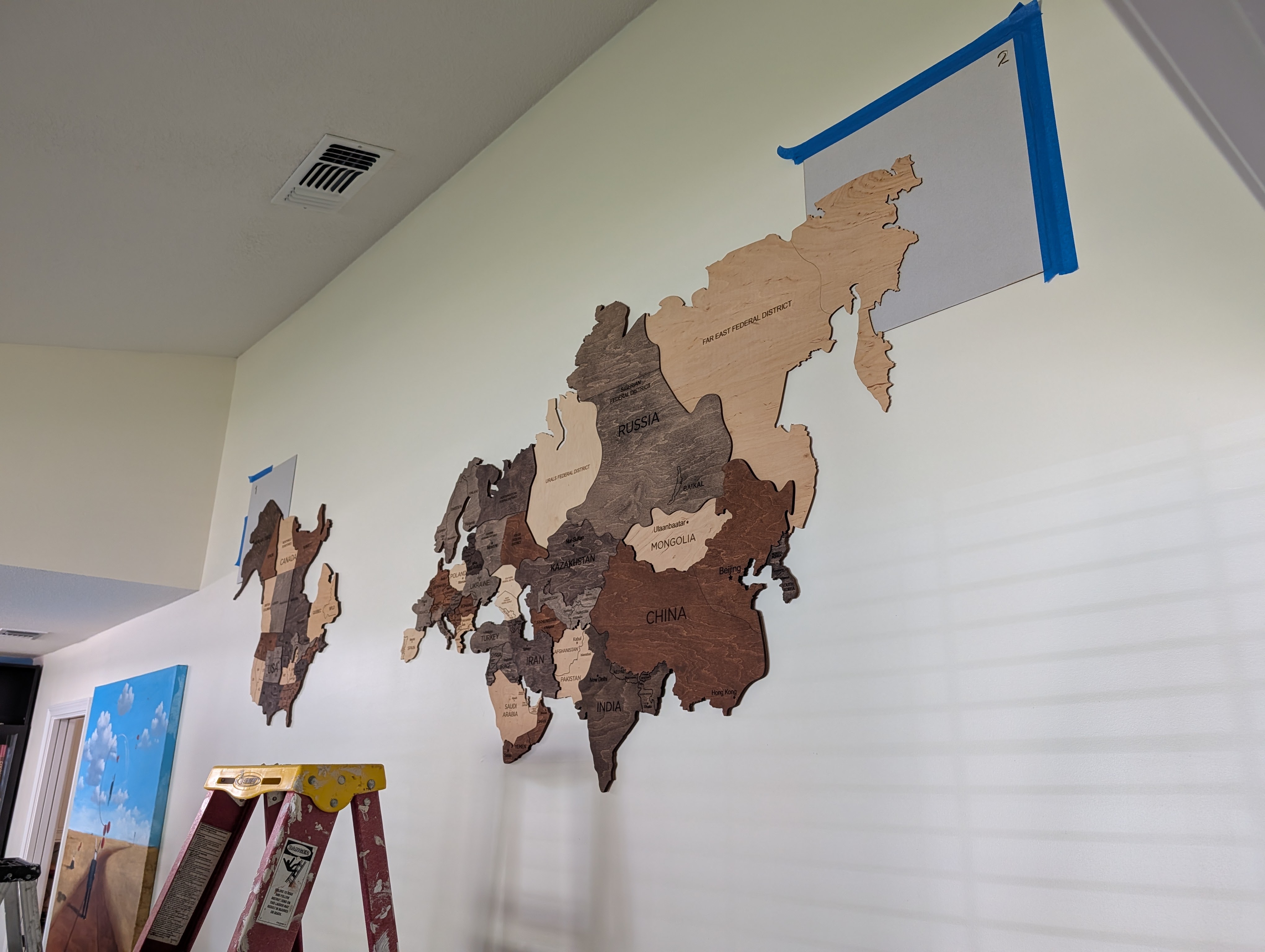 A partially installed wooden world map displayed on a wall, featuring different shades of wood representing various continents and countries, with a piece of paper outlined by blue tape above.