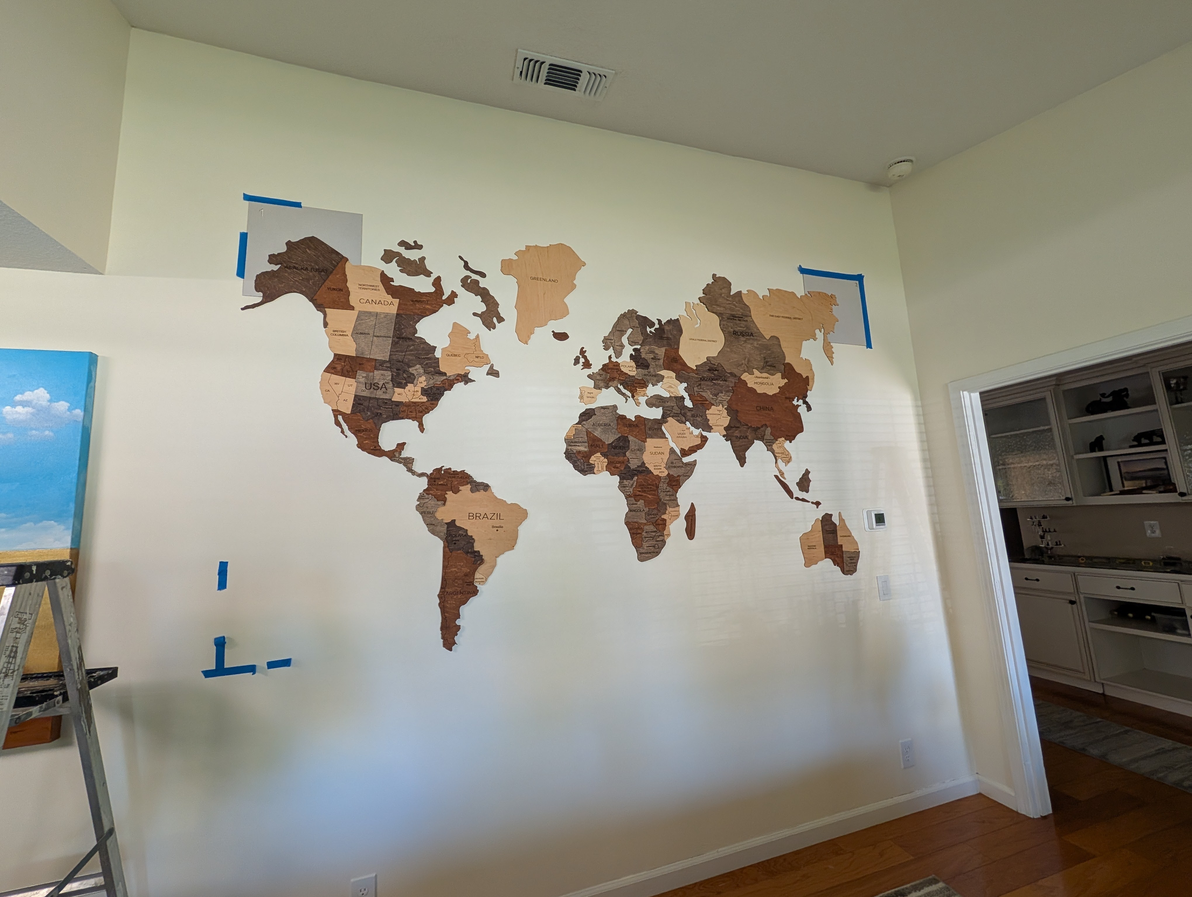 A wooden map of the world is mounted on a wall, featuring various countries in different shades of wood. Blue painter's tape outlines the area for installation, and a painting of a blue sky with clouds is visible on the left side. A ladder is positioned nearby, suggesting an ongoing installation process.