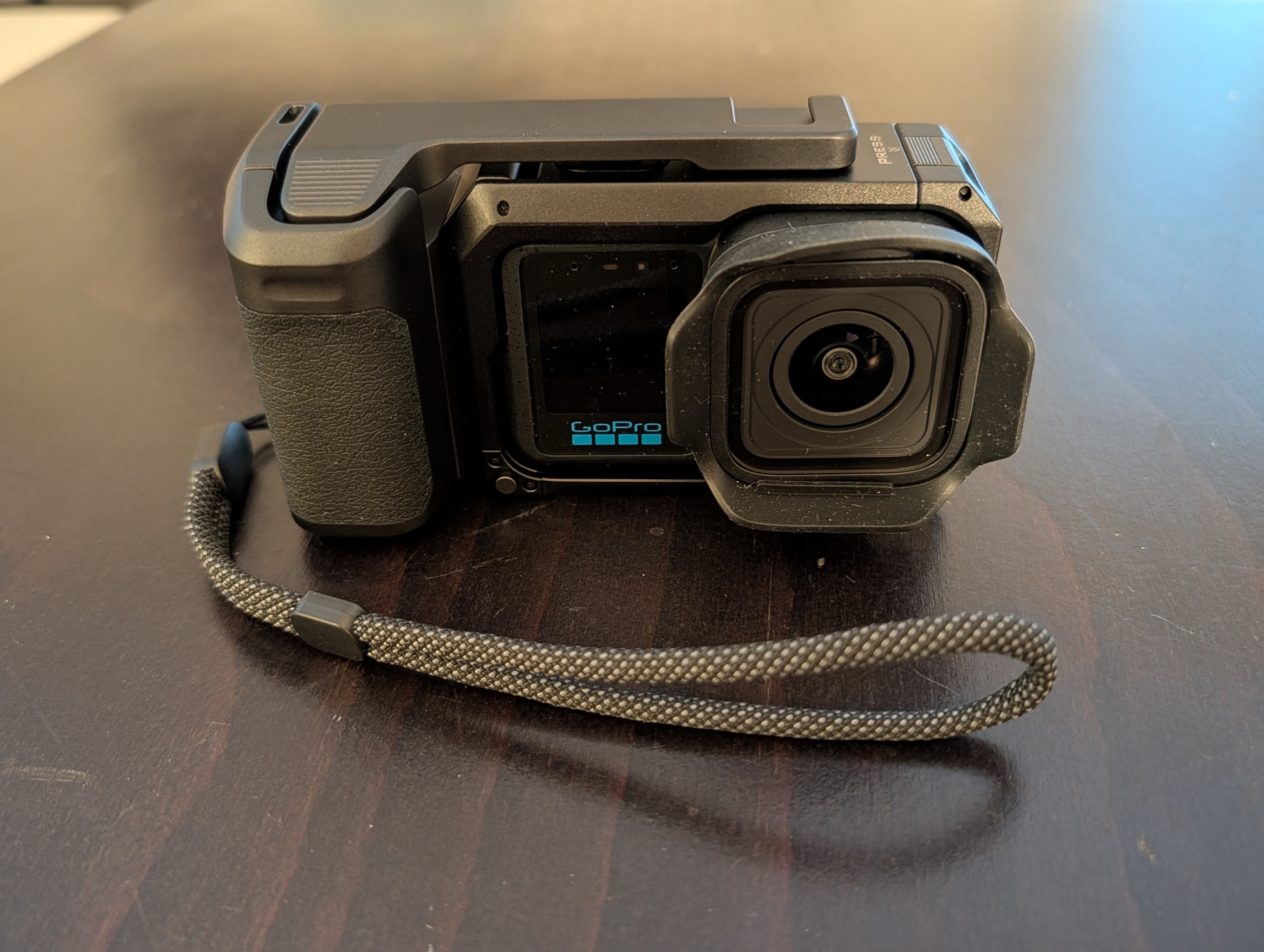 Front view of a GoPro camera with a protective case and strap, resting on a dark wooden surface.