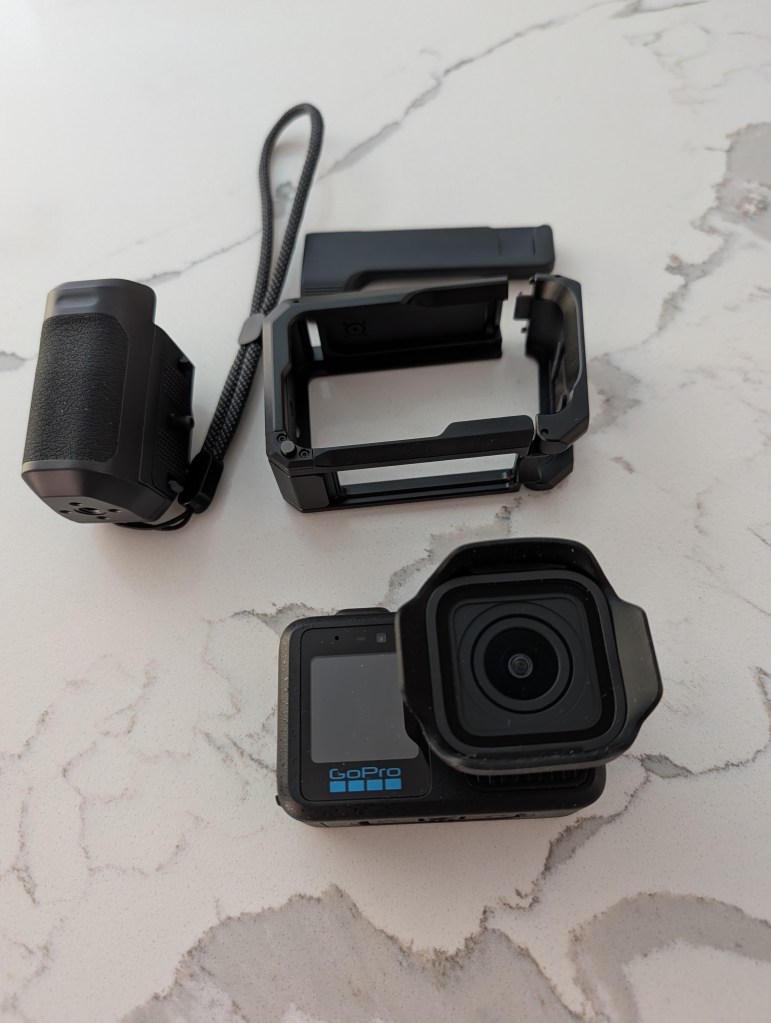 A GoPro camera displayed with an external battery, a protective case, and a strap, placed on a light marble surface.