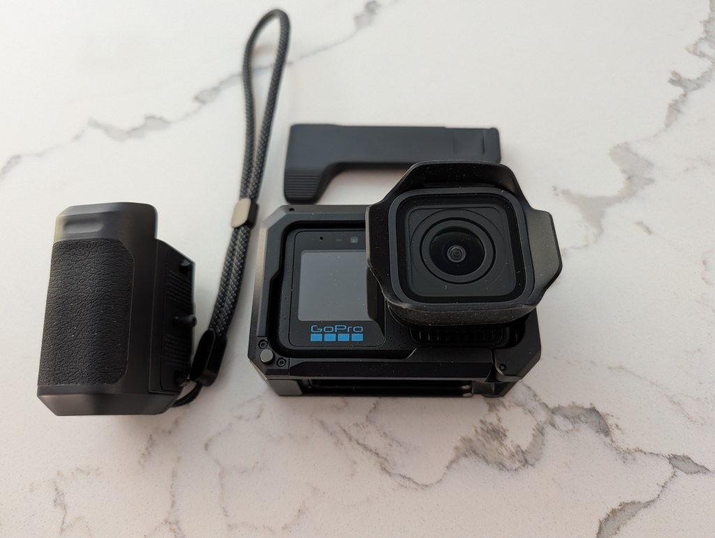 A GoPro camera positioned on a marble surface, featuring a protective lens cover, a display screen, and an attached grip for stabilization.