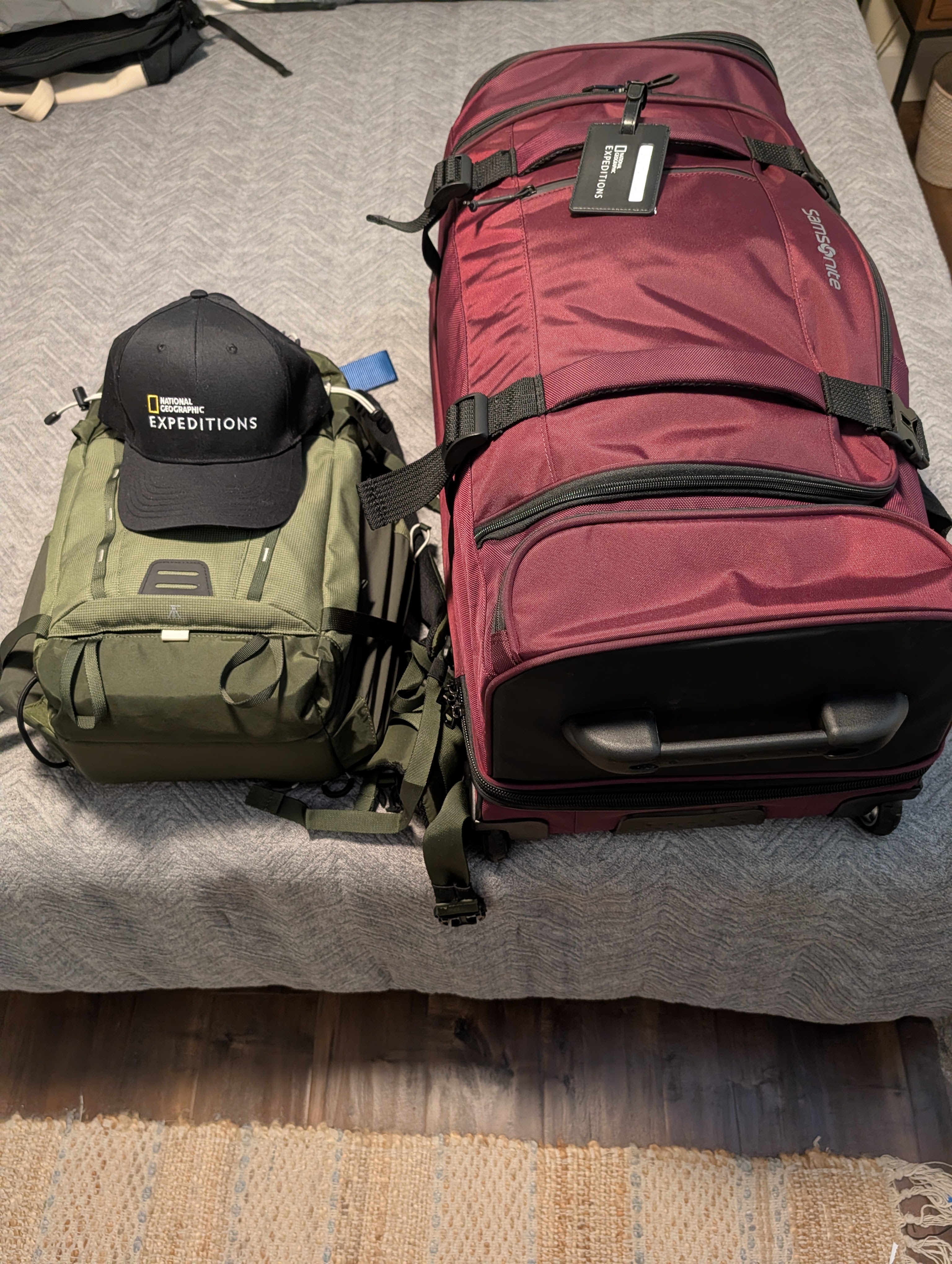 A green backpack and a large maroon travel duffel bag placed on a bed, with a black National Geographic Expeditions cap on top of the backpack.