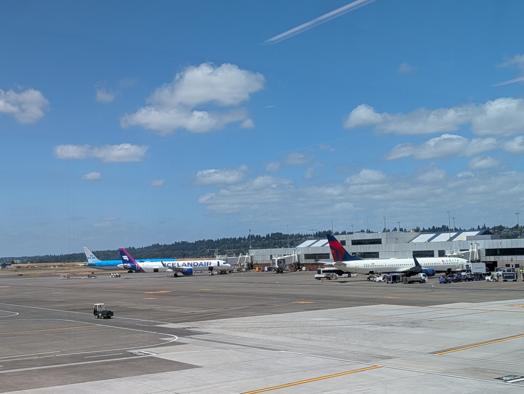 View of an airport runway featuring various aircraft, including a Delta Airlines and Icelandair plane, with a clear blue sky and scattered clouds.