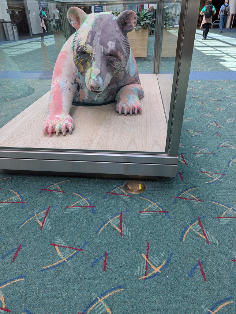 A colorful, artistic bear sculpture displayed in a glass enclosure on a wooden base, with a patterned carpet visible in the foreground.