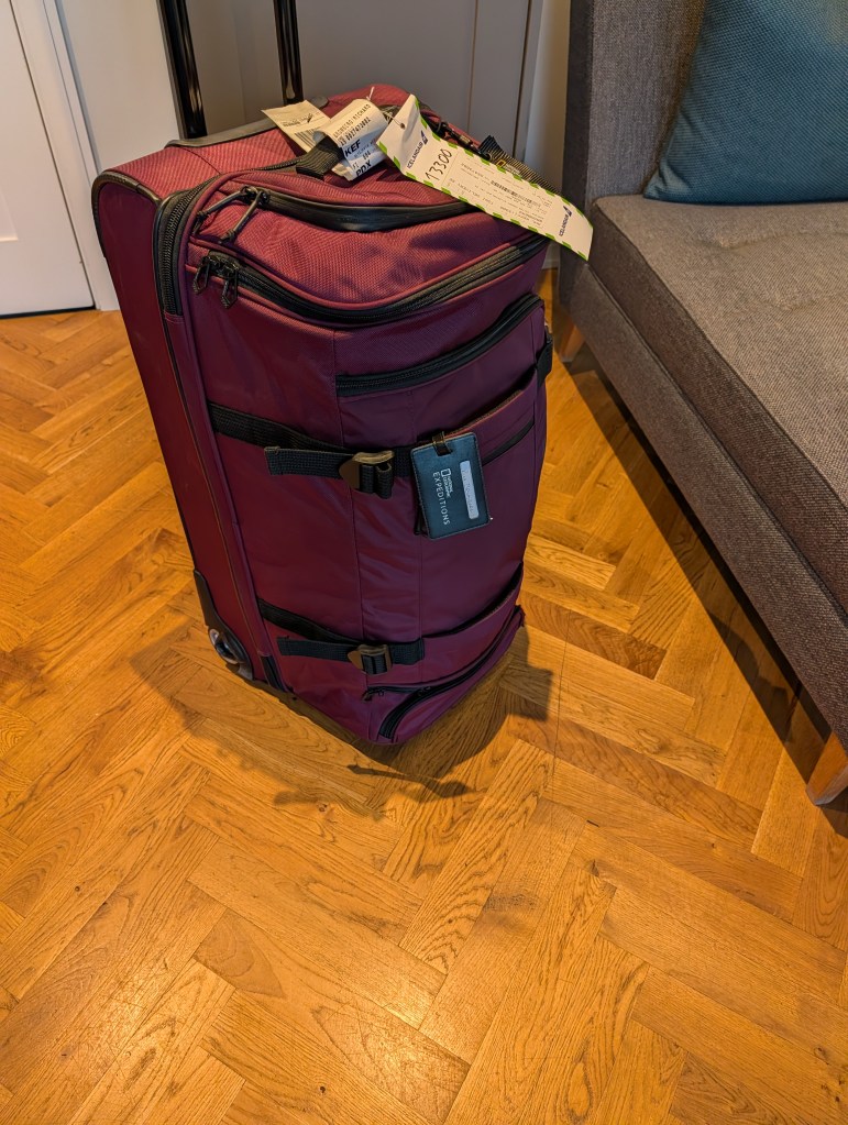 A maroon wheeled suitcase with tags standing on wooden flooring next to a gray armchair.