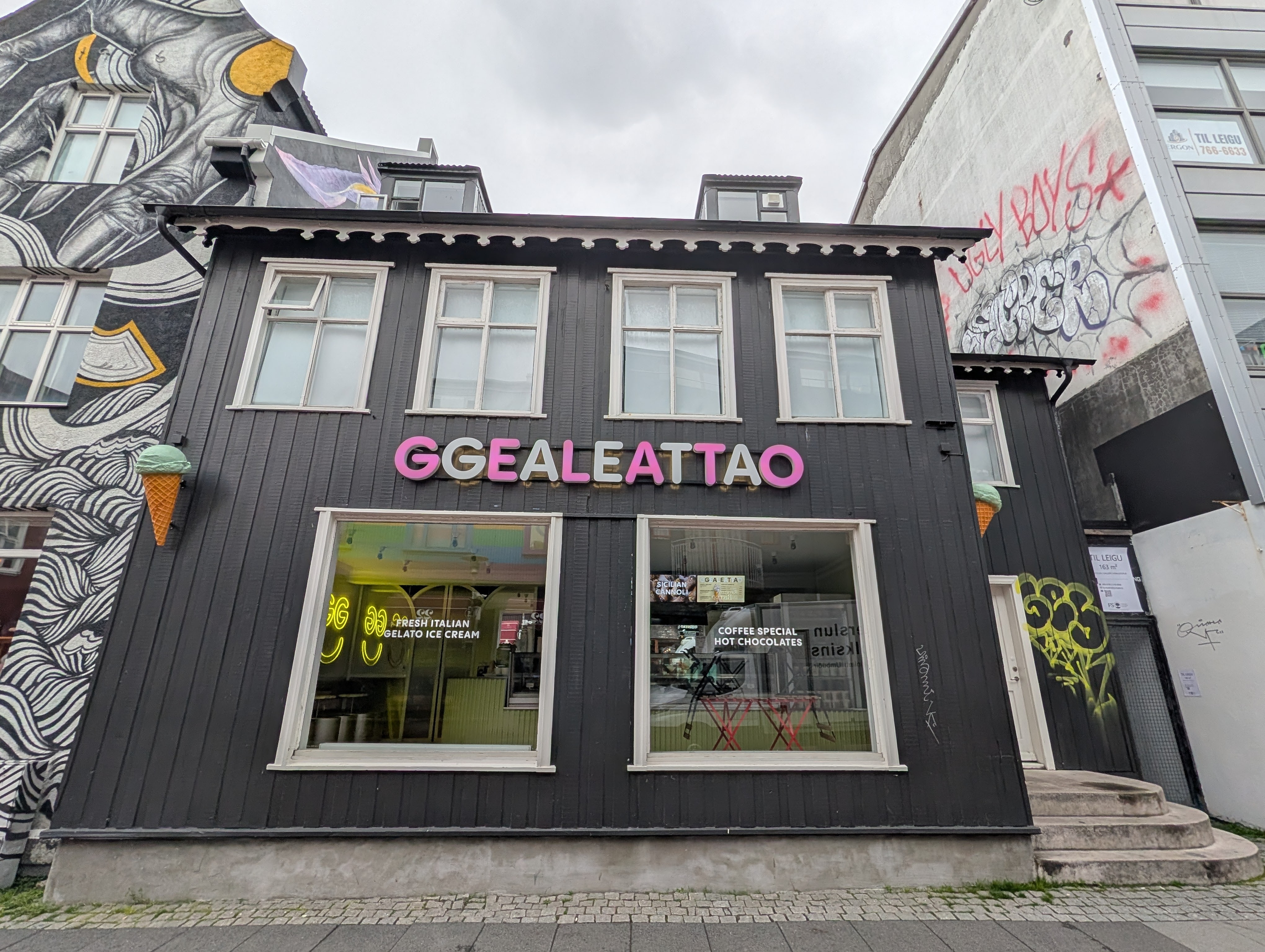 A modern black wooden building featuring large windows and a colorful sign reading 'GGEALEATTAO'. The storefront showcases gelato offerings and has an ice cream cone decoration on the wall. Nearby, there is street art on adjacent buildings.