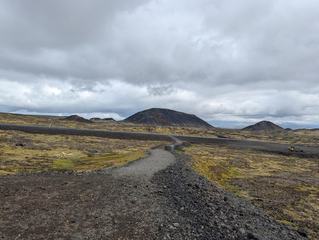 A winding gravel path through a rocky landscape with volcanic hills in the background under a cloudy sky.