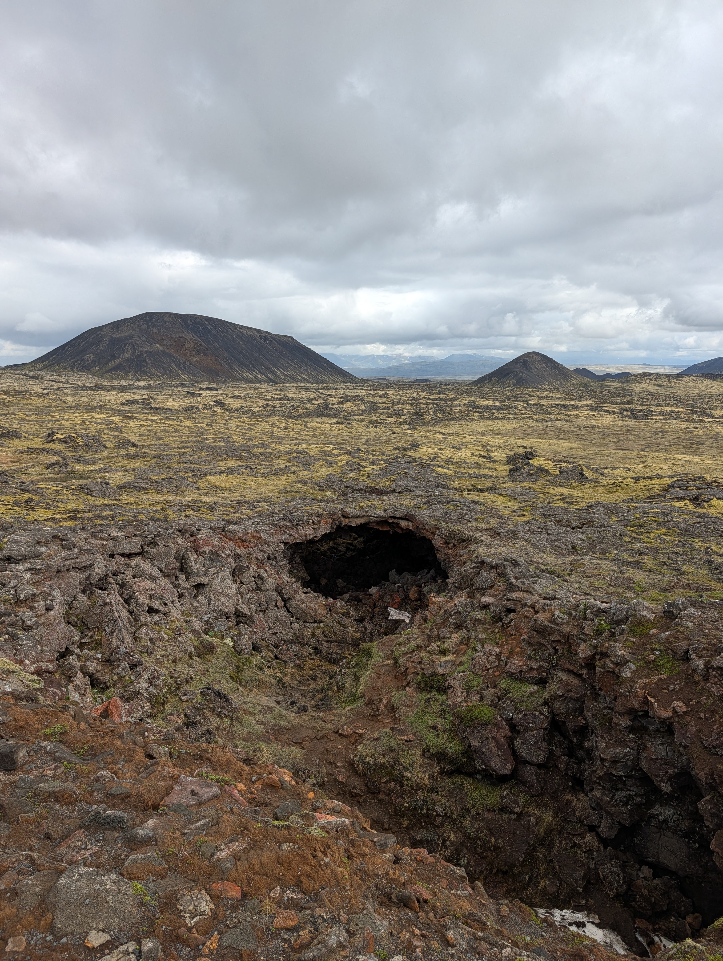 A view of rugged volcanic terrain with a prominent lava tube entrance in the foreground, surrounded by rocky ground and distant volcanic hills under a cloudy sky.