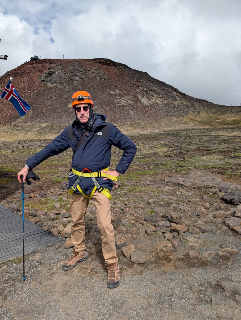 A person standing in front of a volcanic landscape, wearing a dark jacket, an orange helmet, and a safety harness. They hold a trekking pole and pose confidently with one hand on their hip. In the background, a rocky hill and an Icelandic flag are visible.