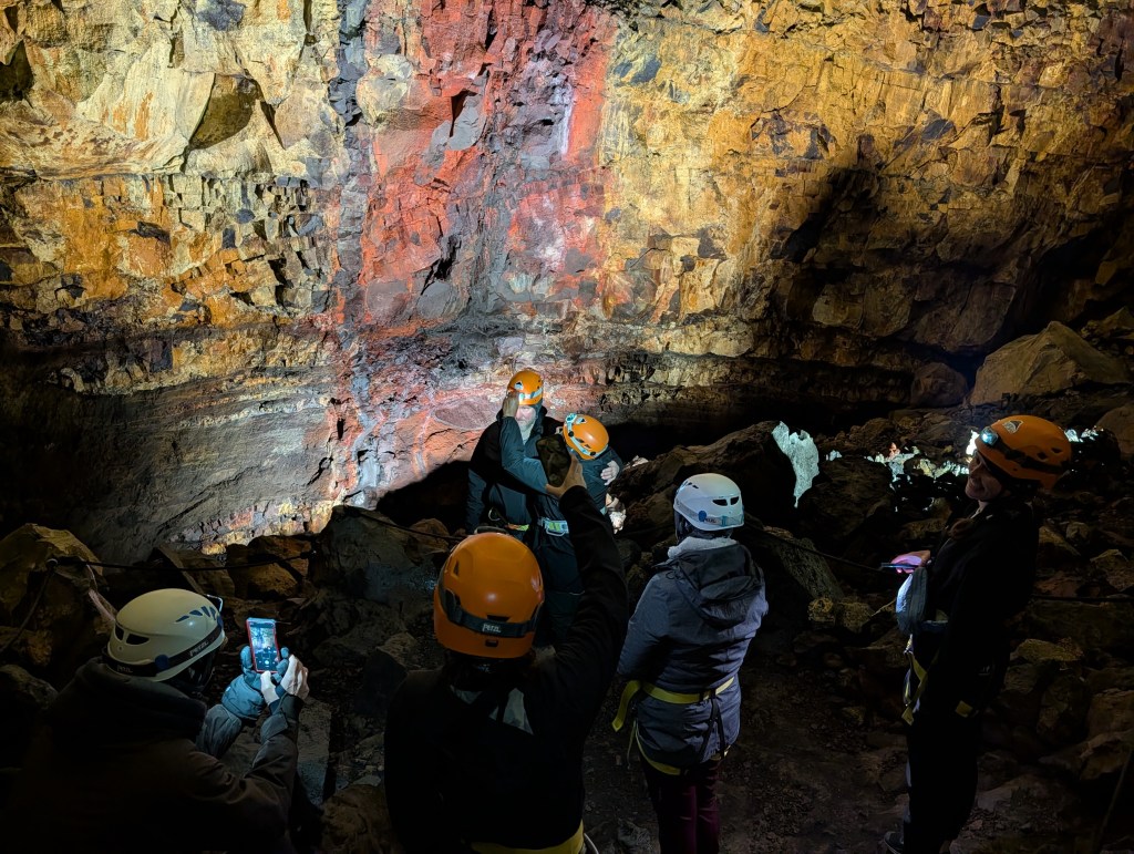 A group of six individuals wearing helmets and headlamps inside a dark cave, exploring the colorful rock formations illuminated by artificial lights.