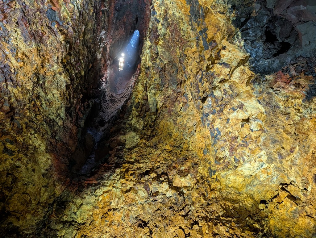 An aerial view of a rocky cave with illuminated areas and textured orange and yellow walls, revealing mineral deposits and a narrow passageway.