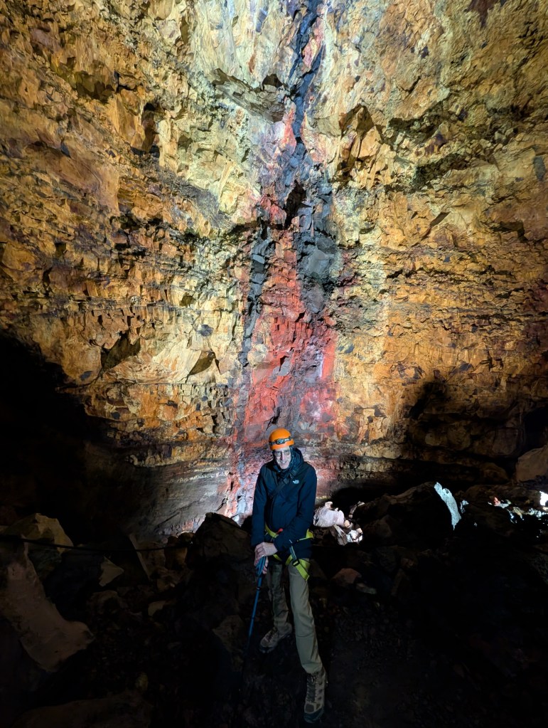 A climber stands in a colorful cave, illuminated by artificial light, showcasing textured rock formations in shades of orange, brown, and black.