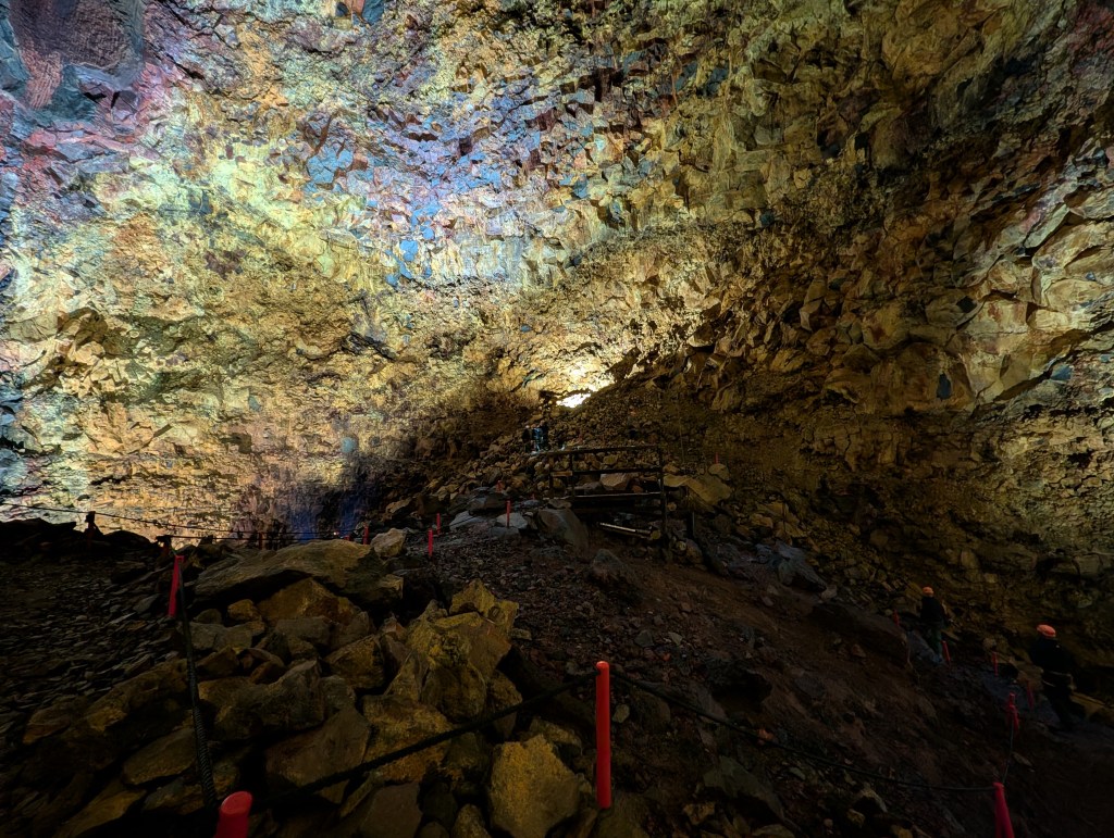 A colorful cave interior with a textured ceiling and rocky ground. Small figures can be seen exploring the area, with safety barriers marked by red posts.