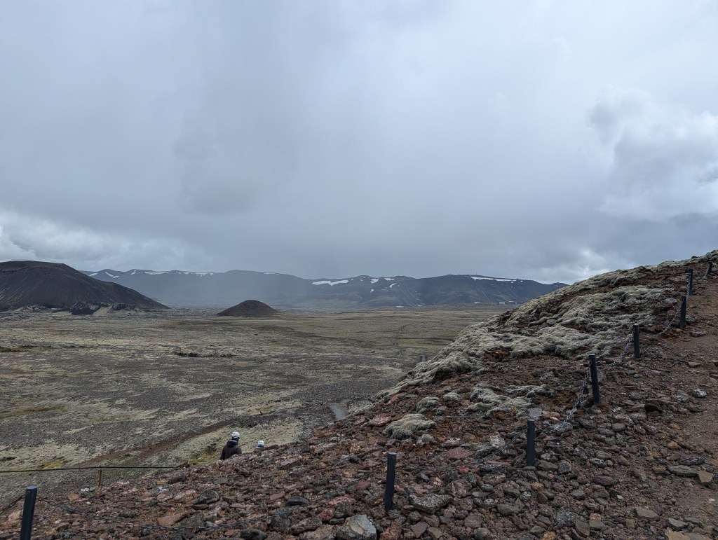A panoramic view of a volcanic landscape featuring mountains and a cloudy sky, with two individuals walking on a rocky path in the foreground.