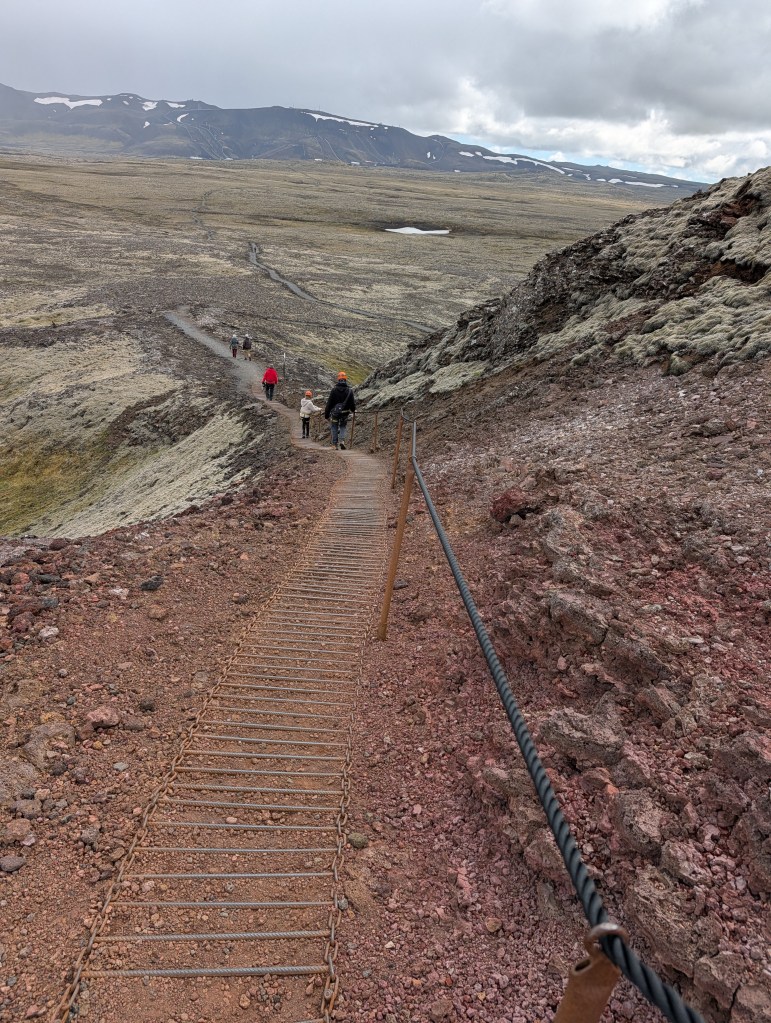 A hiking trail with a metal railing leads down a rocky and rugged landscape, where several people are walking in the distance under a cloudy sky.