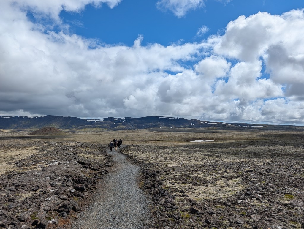 A group of hikers walking along a rocky path in a vast, open landscape with mountains and clouds in the background.