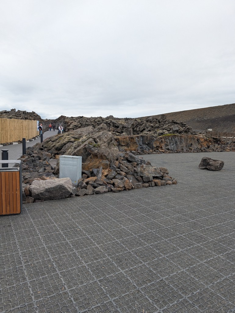 A rocky landscape with a walkway, featuring a wooden fence and a group of people in the background. Large volcanic rocks are scattered around, with a waste bin and a utility box visible.