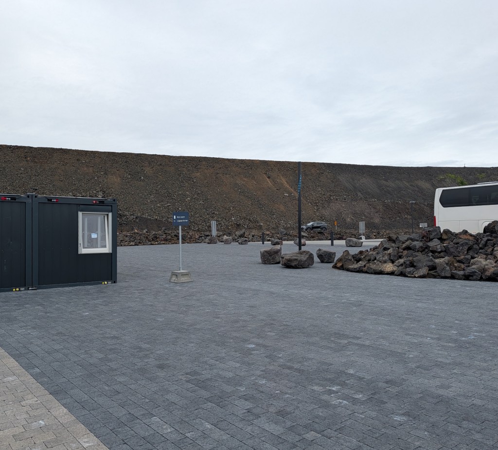 An empty parking area with gray paving stones, featuring black shipping containers, signposts, and a rocky hillside in the background under a cloudy sky.