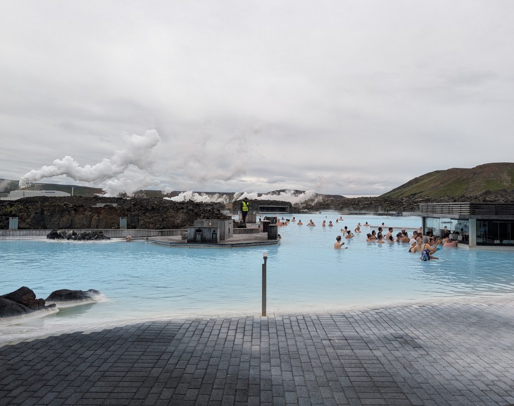 A geothermal spa with people relaxing in blue water, surrounded by rocky terrain and steam rising from the landscape.