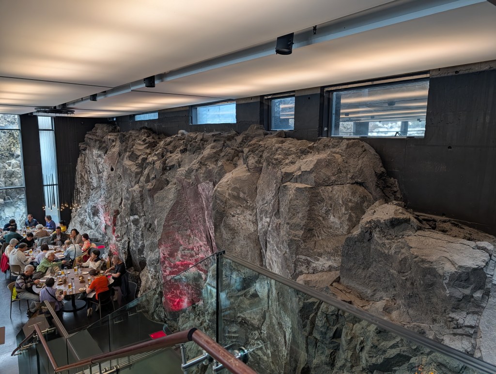 Interior of a restaurant featuring a large natural rock wall, with diners seated at tables below and large windows providing natural light.