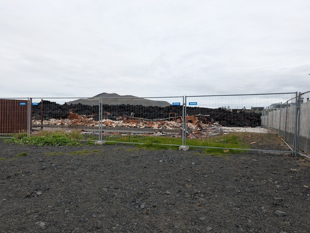 A construction site with a fenced area containing piles of debris and rubble, set against a cloudy sky and a distant hill.