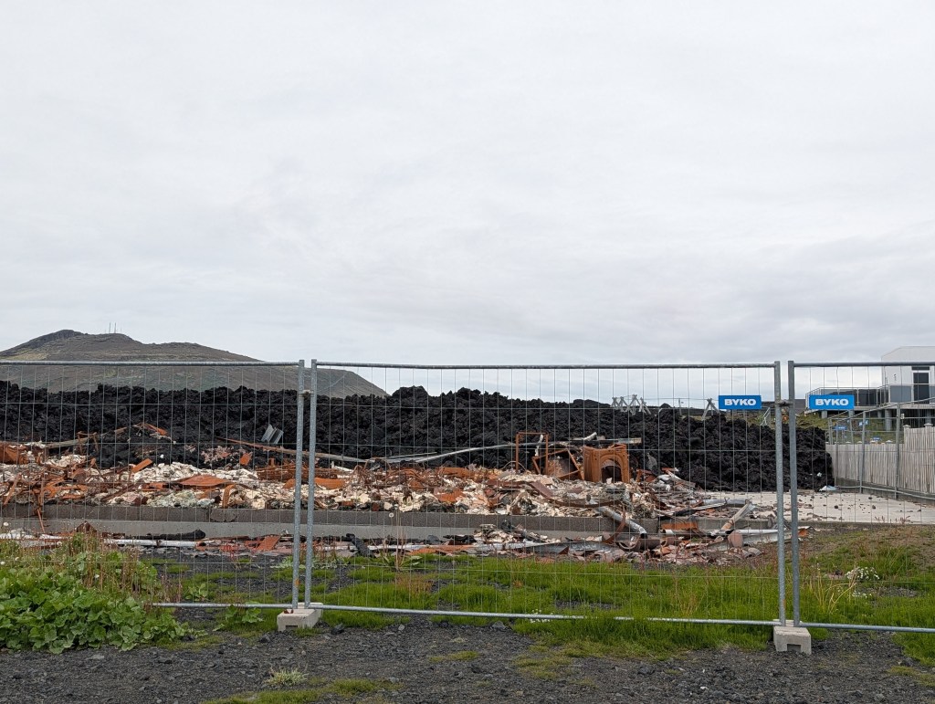 View of a demolished structure surrounded by a fence, with debris and rubble in the foreground and a hillside in the background under a cloudy sky.